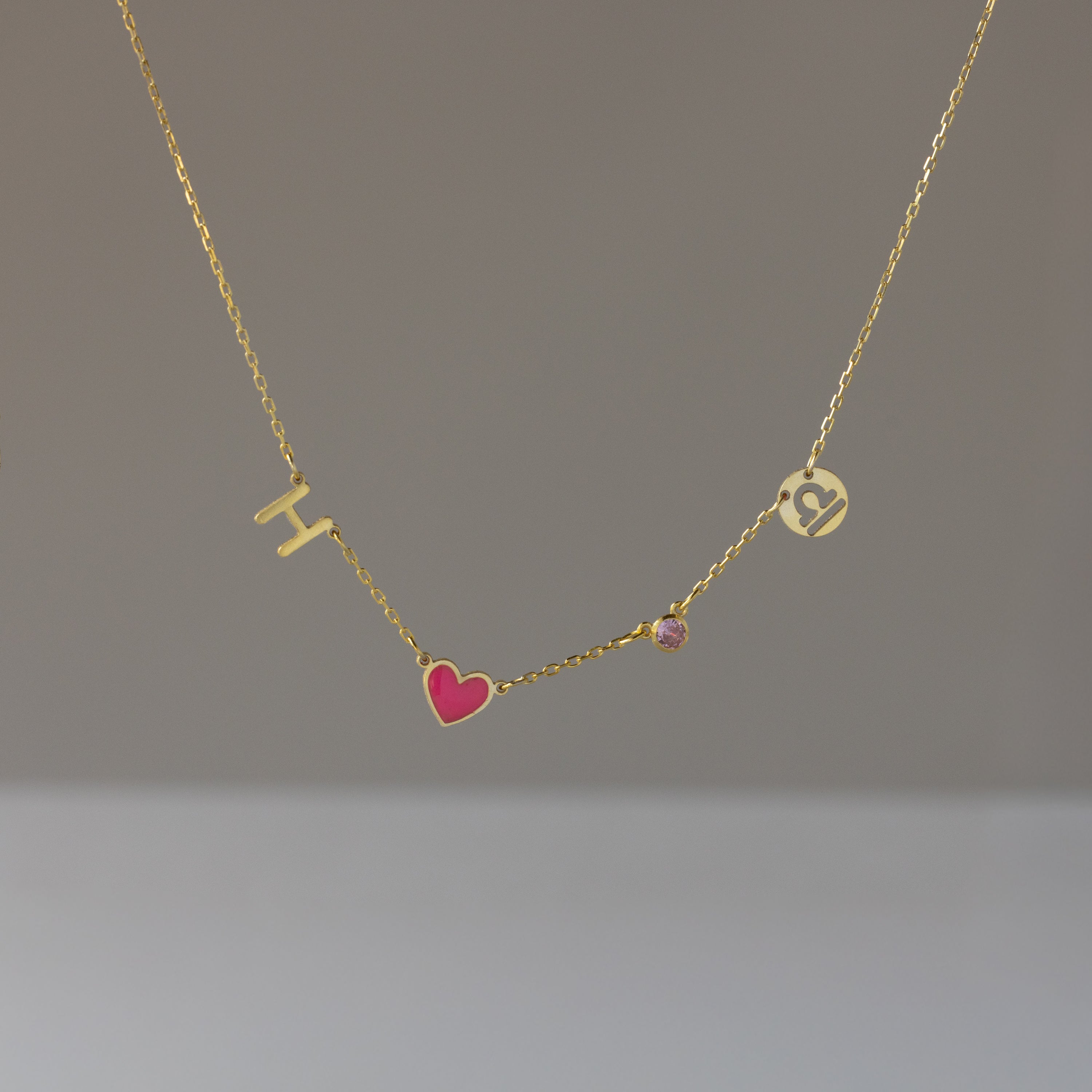 The Dainty Space Charm Necklace features a gold chain with an H charm, pink heart, small pink gem, and a round pendant with a C cutout—perfect for adding customizable elegance to your everyday style.