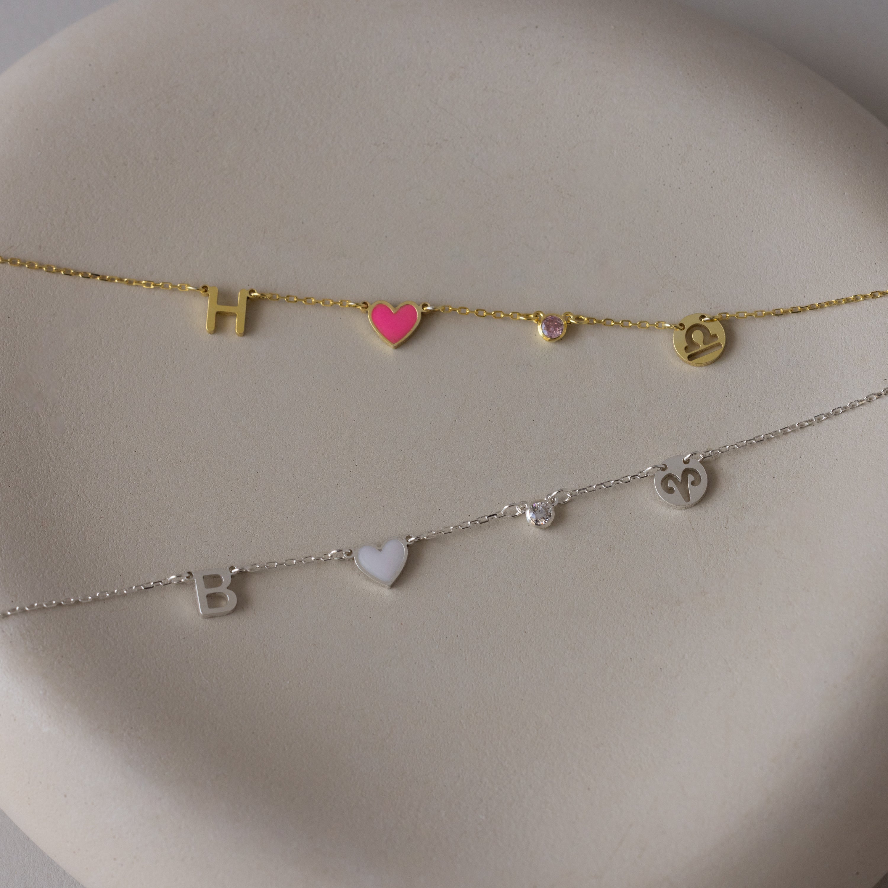 Two delicate bracelets with letter, heart, and symbol charms—one gold-toned, one silver-toned—on a cream surface; perfect for pairing with the Dainty Space Charm Necklace or stacking alongside your favorite birthstone necklace.
