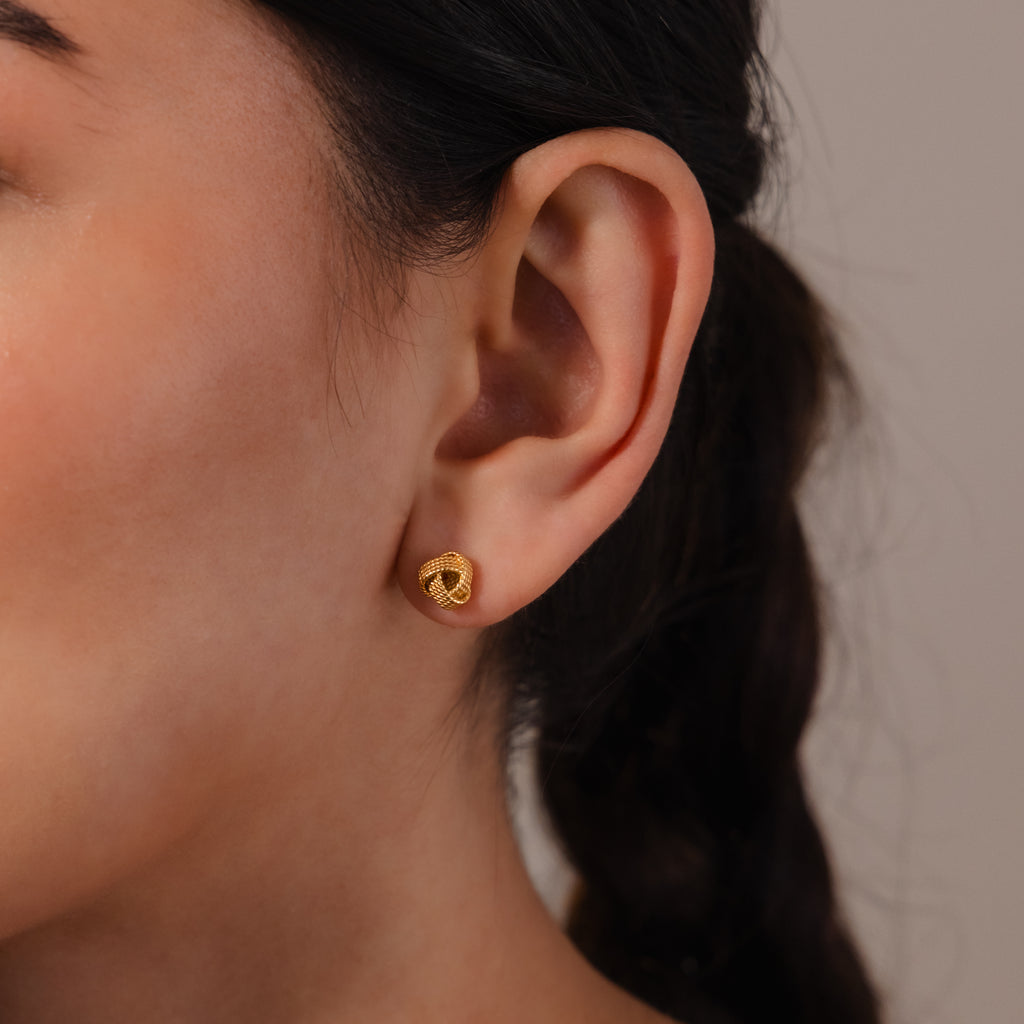 Close-up of a woman's ear wearing Rope Knot Studs, small round gold earrings; her hair is styled in a braid for an elegant, classic look.