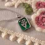 The Emerald Birth Flower Charm Necklace features a green enamel floral design and shines against cream and pink embroidered fabric, making it a charming vintage-inspired addition to any jewelry collection.
