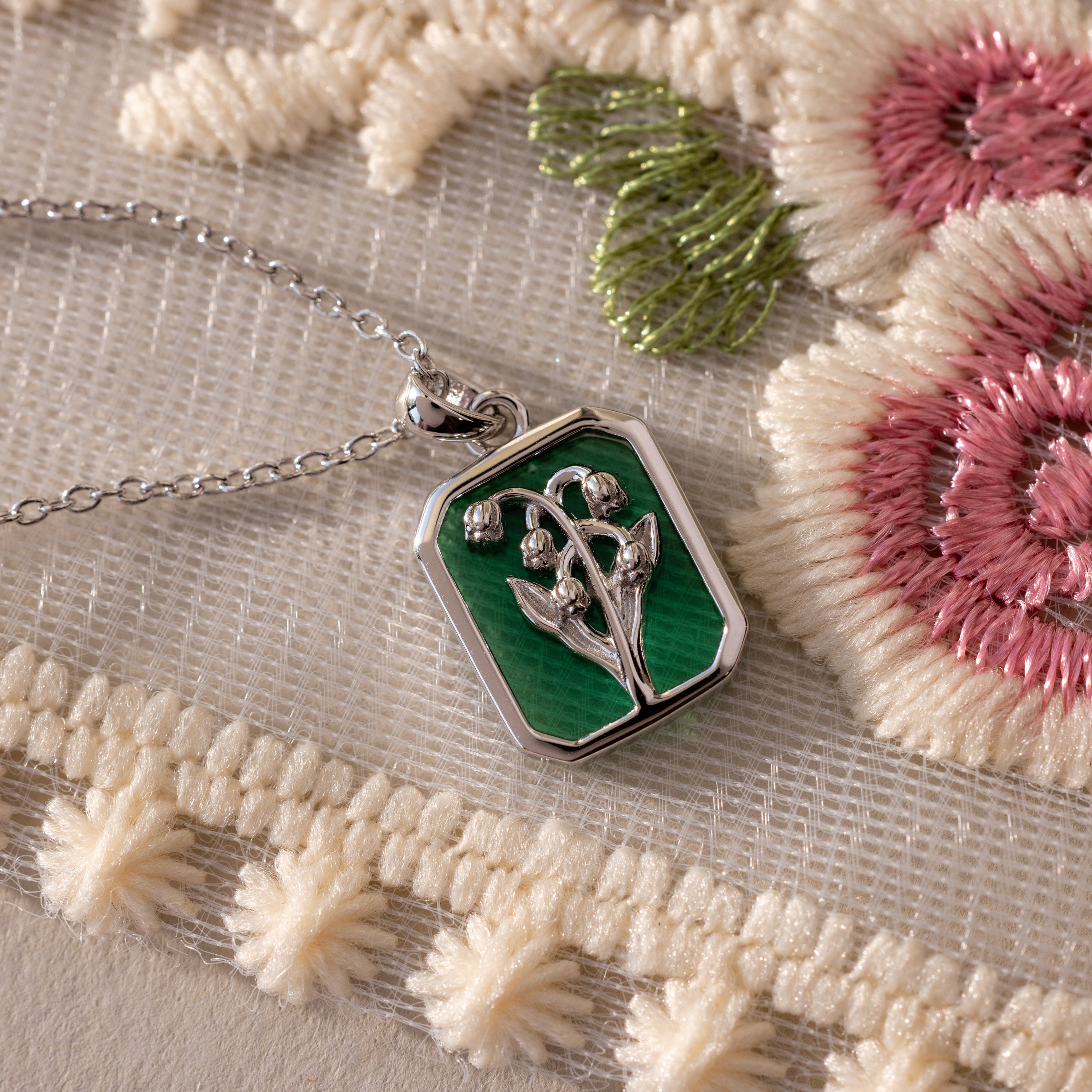 The Emerald Birth Flower Charm Necklace features a green enamel floral design and shines against cream and pink embroidered fabric, making it a charming vintage-inspired addition to any jewelry collection.