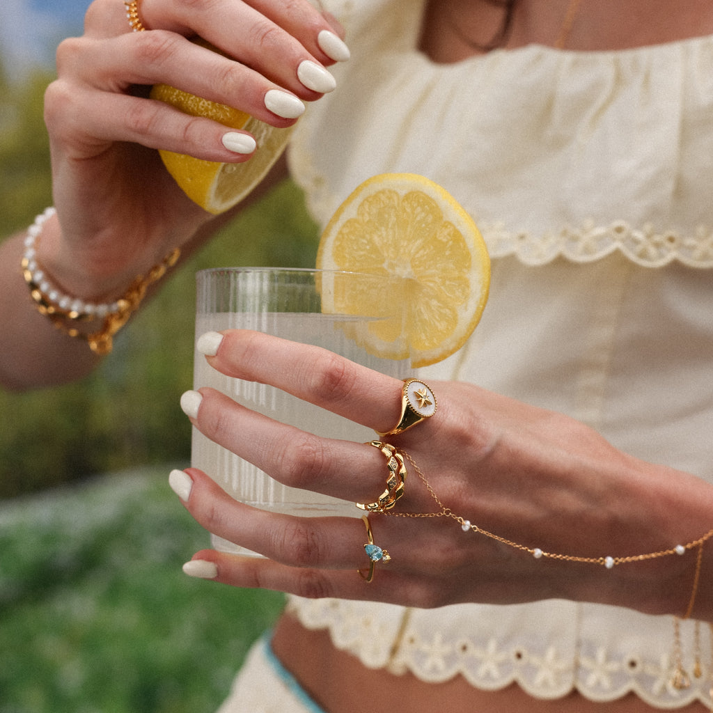 Woman wearing the North Star Signet Ring, gold rings—including a pearl ring—and bracelets, in a cream ruffled top, holding a lemon and a glass of water.