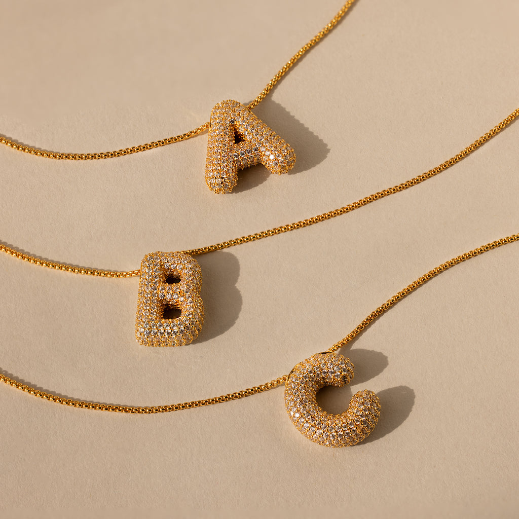 Three Pave Puff Letter Necklaces in 18K Gold feature crystal-studded A, B, and C pendants, elegantly displayed on a beige background.