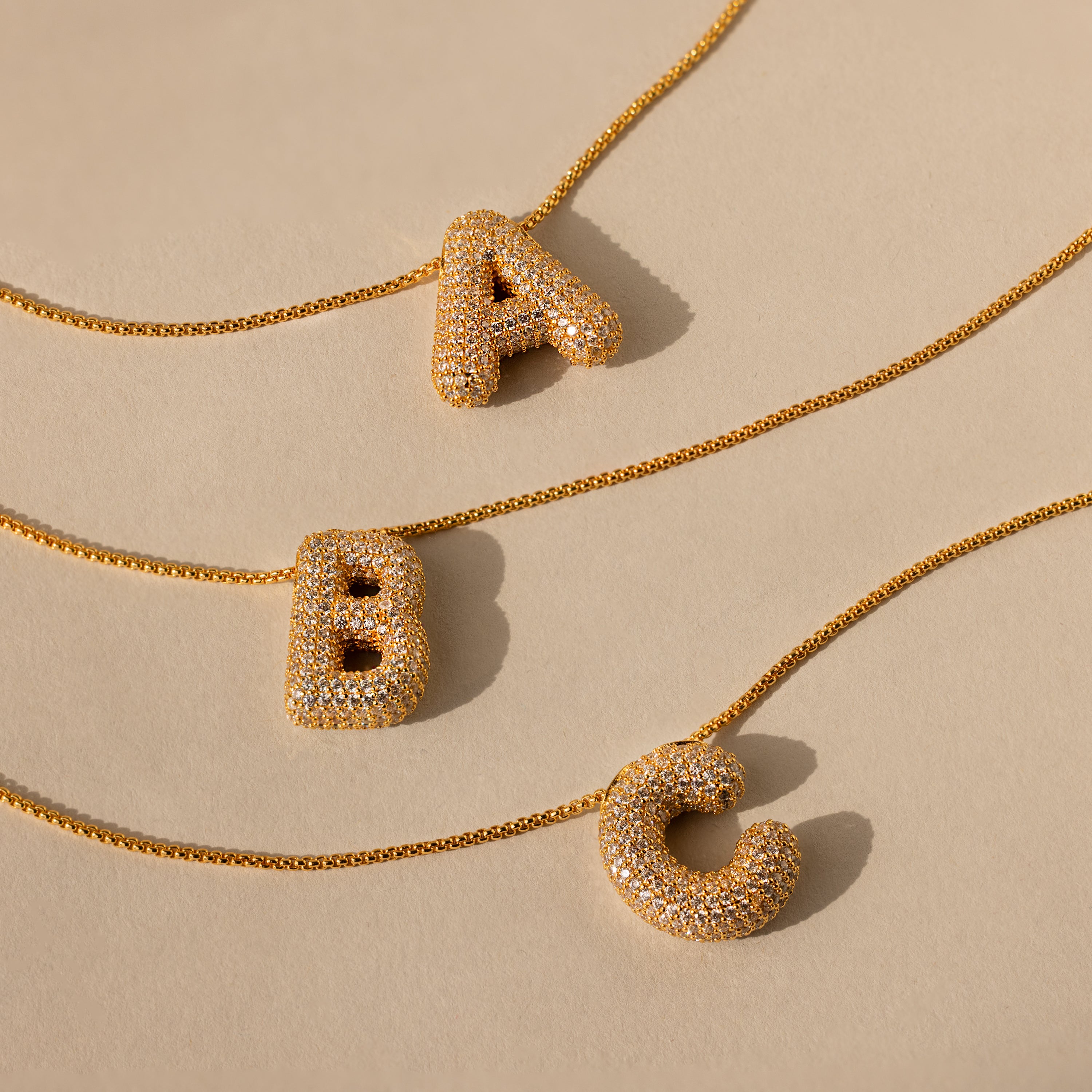 Three Pave Puff Letter Necklaces in 18K Gold feature crystal-studded A, B, and C pendants, elegantly displayed on a beige background.