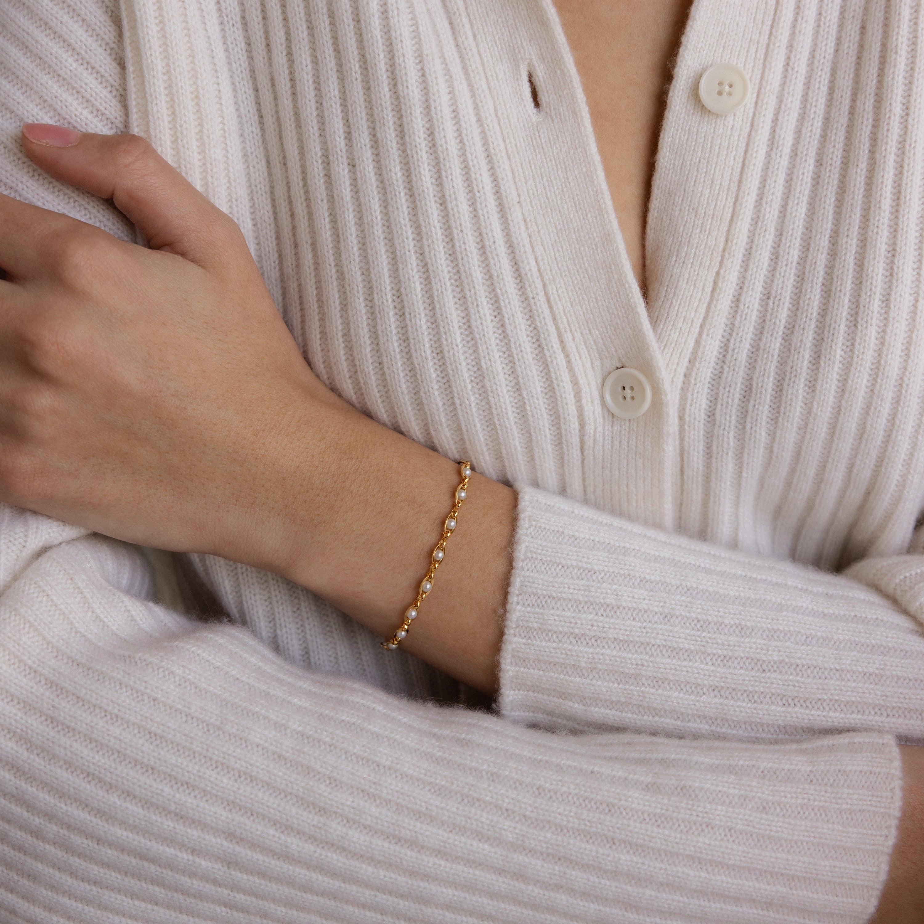 A person in a cream ribbed sweater wears the Eternity Pearl Bracelet in 18K Gold, pairing it with an infinity chain for an elegant jewelry look.