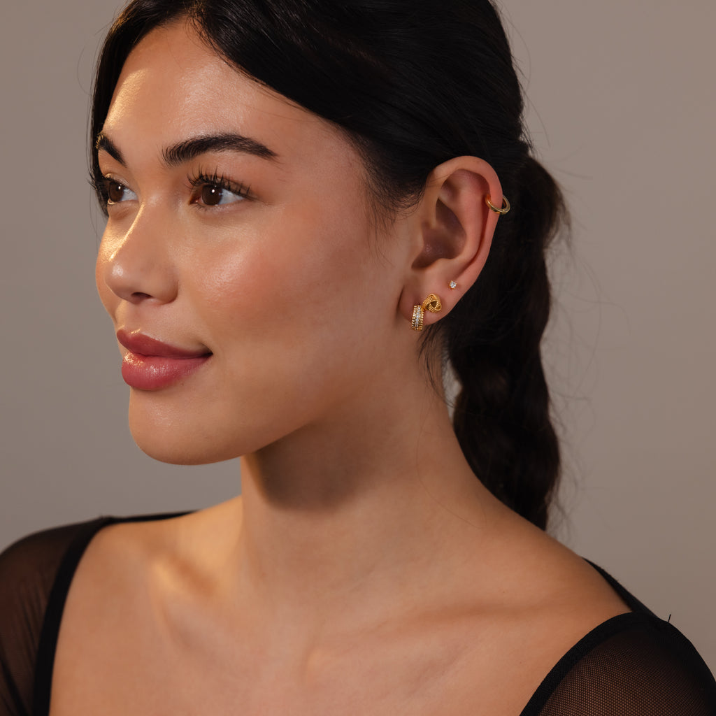 Woman with braided dark hair, wearing Rope Knot Studs in 18K Gold and a sheer black top, smiles softly as she gazes to the left.