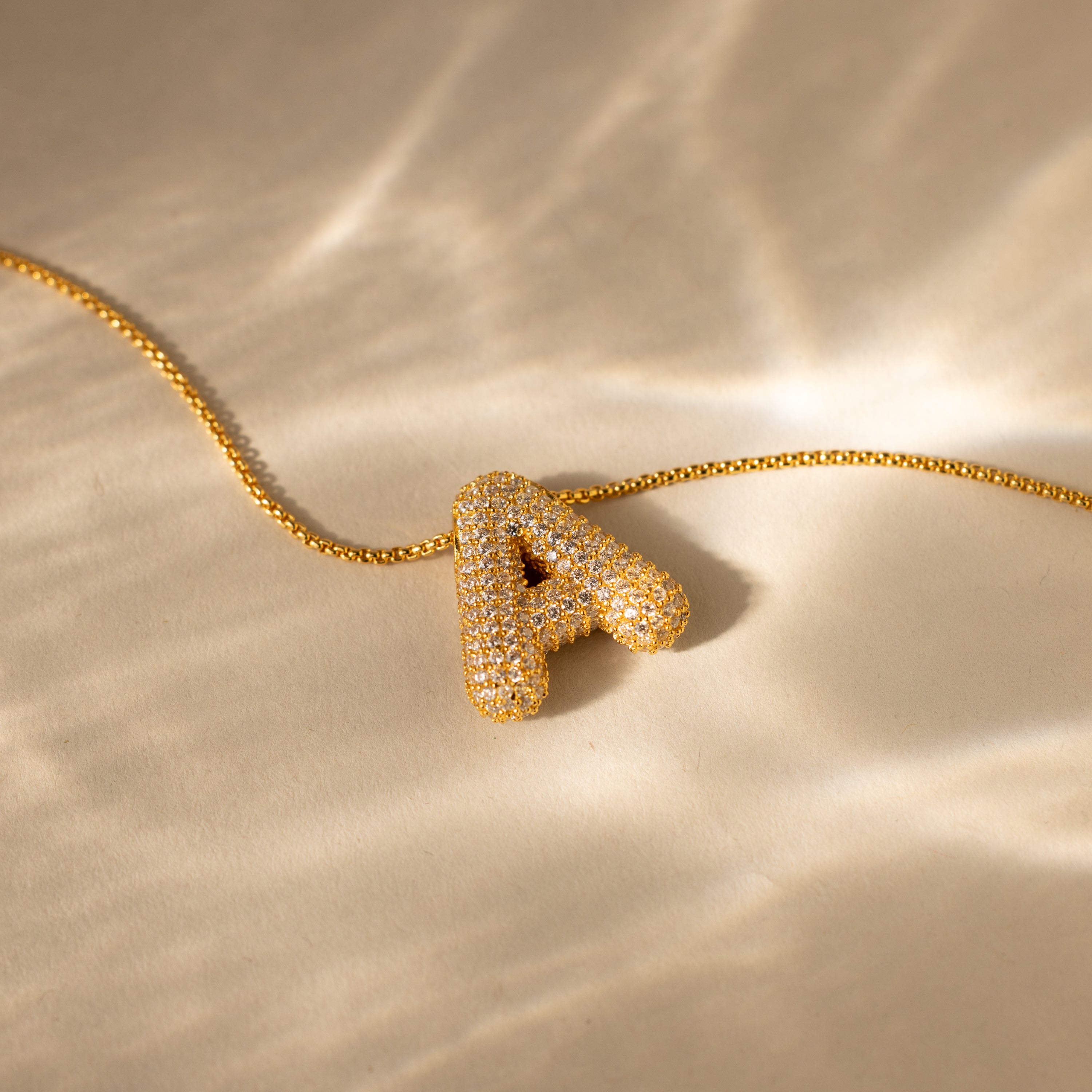 The Pave Puff Letter Necklace in 18K Gold features a jeweled letter A pendant on a gold chain, sparkling on a beige surface—combining a stunning gold letter charm with elegant pave diamonds.