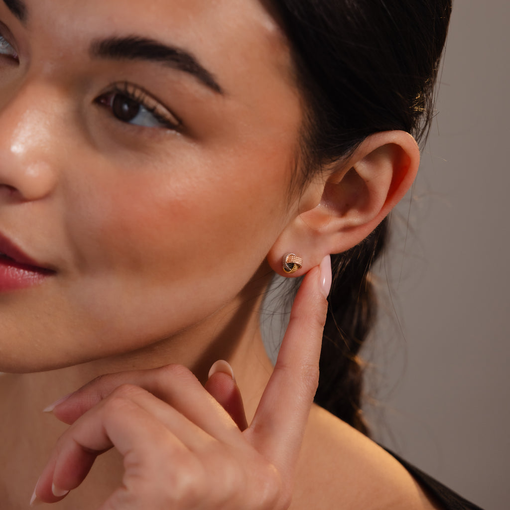 A woman with braided hair smiles softly while pointing to a Rope Knot Stud earring on her ear.