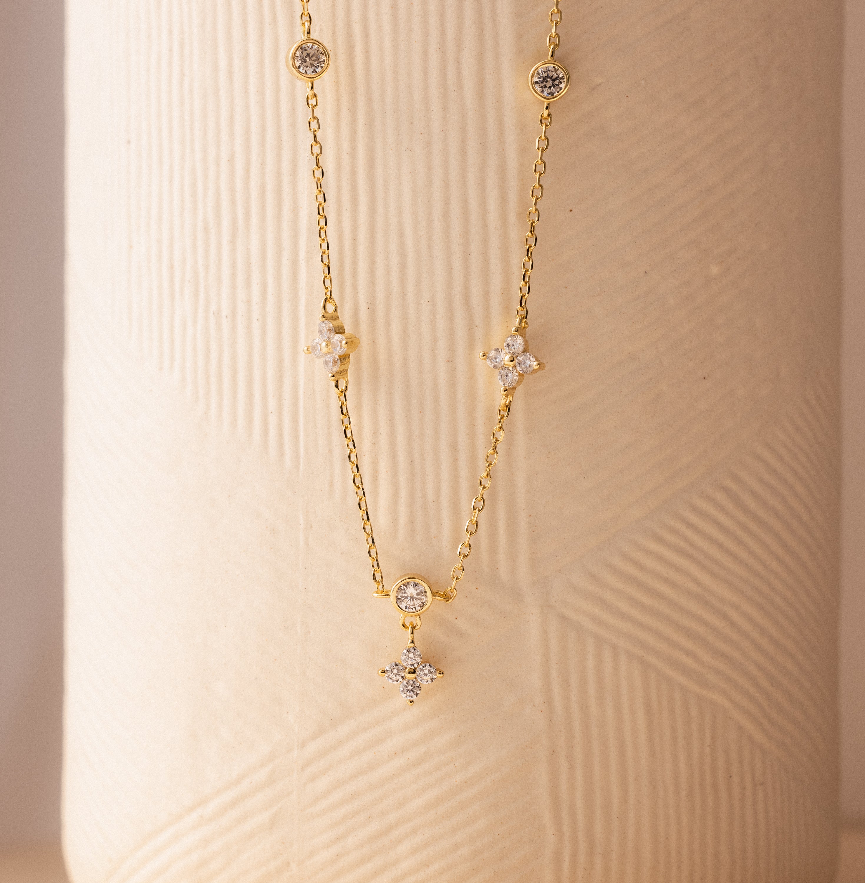 The Ivy Diamond Station Necklace features diamond-shaped and round gemstone accents, evoking a floral design, displayed on a textured beige surface.