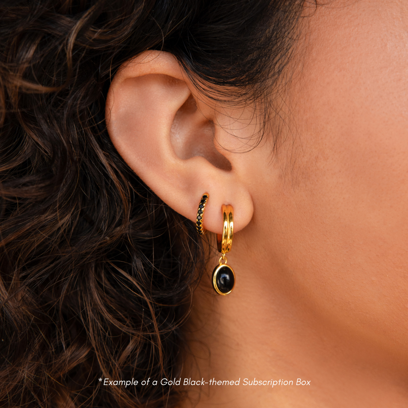 Close-up of a woman's ear with two 18K Gold hoops—one featuring a black stone, the other with green accents. Get inspired for your next pair from the Monthly Earrings Subscription Box.