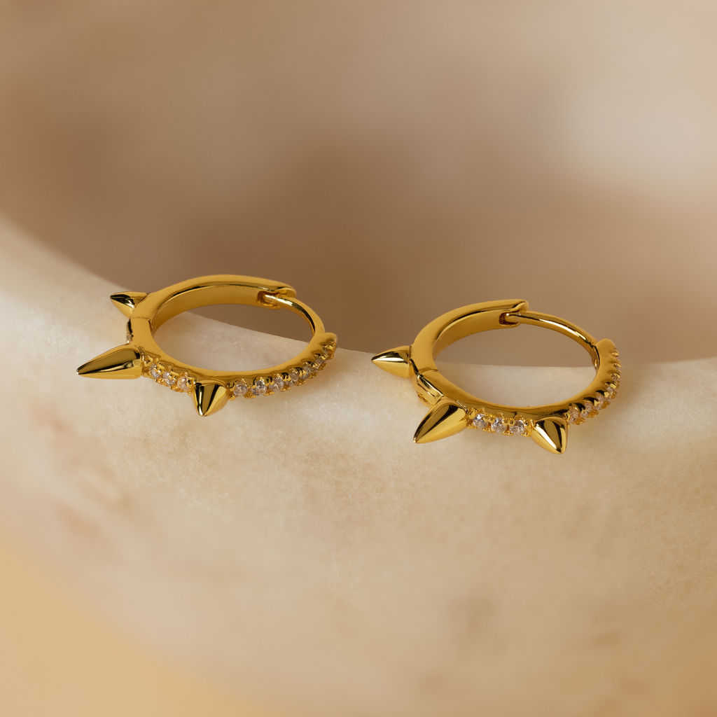 Pave Triple Spike Huggies with sparkling stones offer a bold, minimalist look as they rest elegantly on a smooth beige surface.