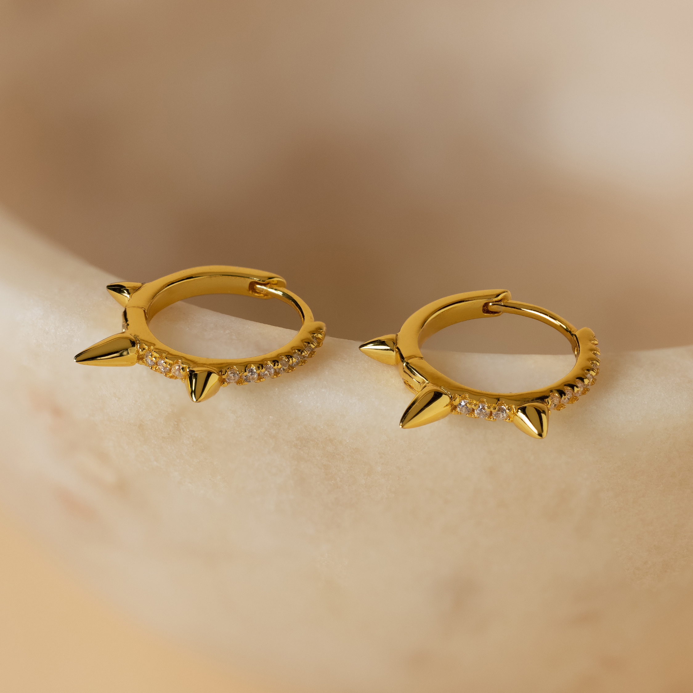 Pave Triple Spike Huggies with sparkling stones offer a bold, minimalist look as they rest elegantly on a smooth beige surface.