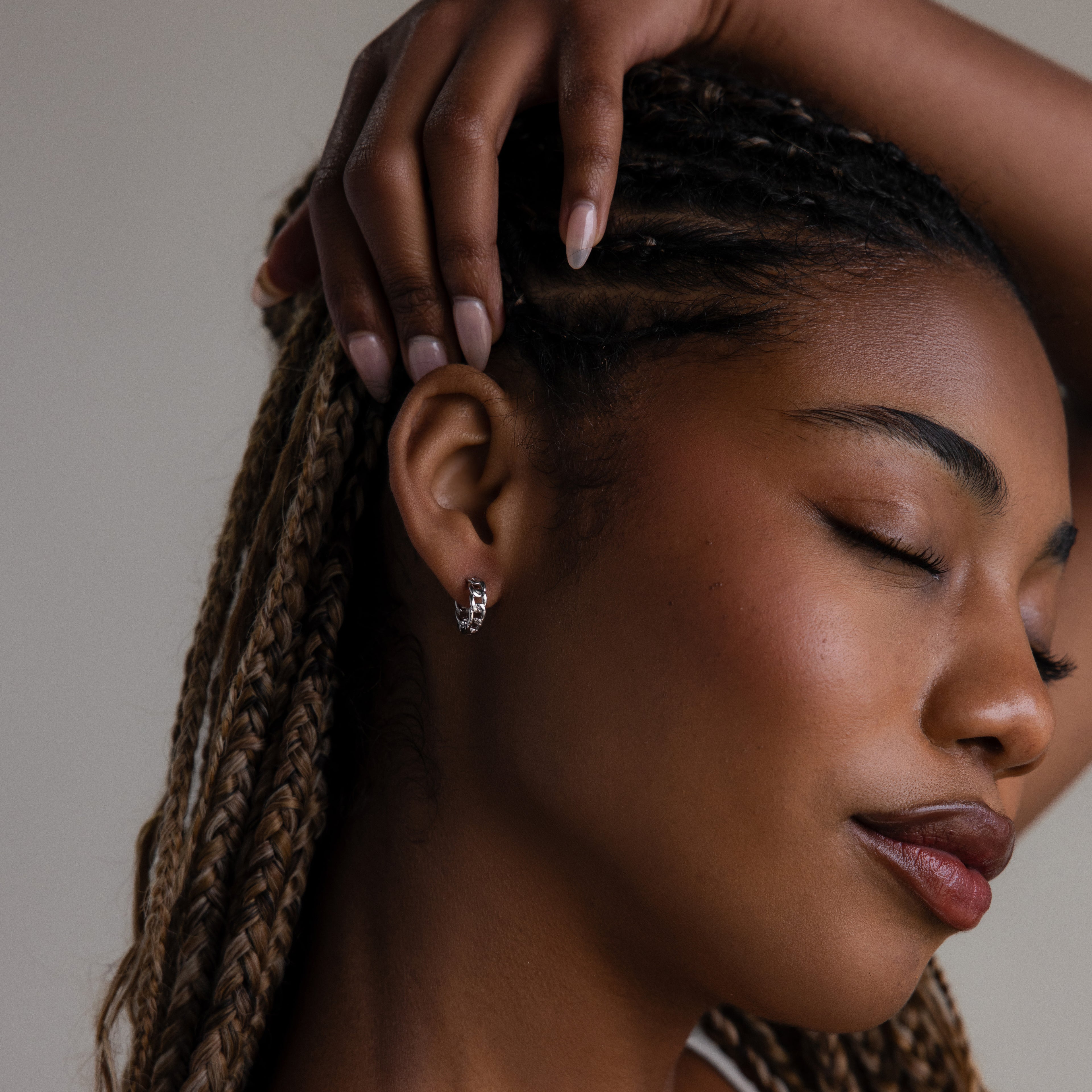 Close-up of a woman with braided hair and silver hoop earrings and her hand gently touching her head against a neutral background