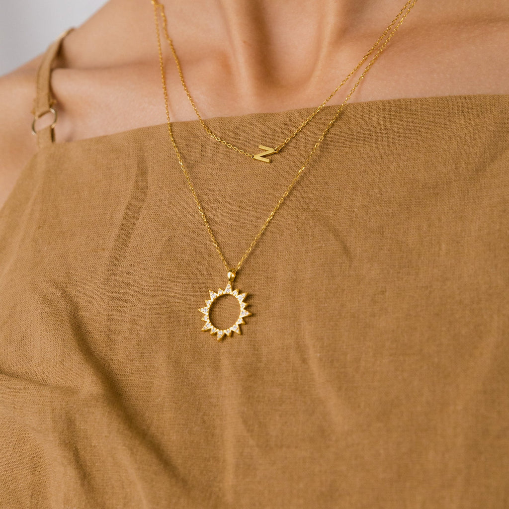 Close-up of a person wearing layered gold necklaces, featuring the Pave Sun Necklace with sparkling pave accents and a letter N charm.