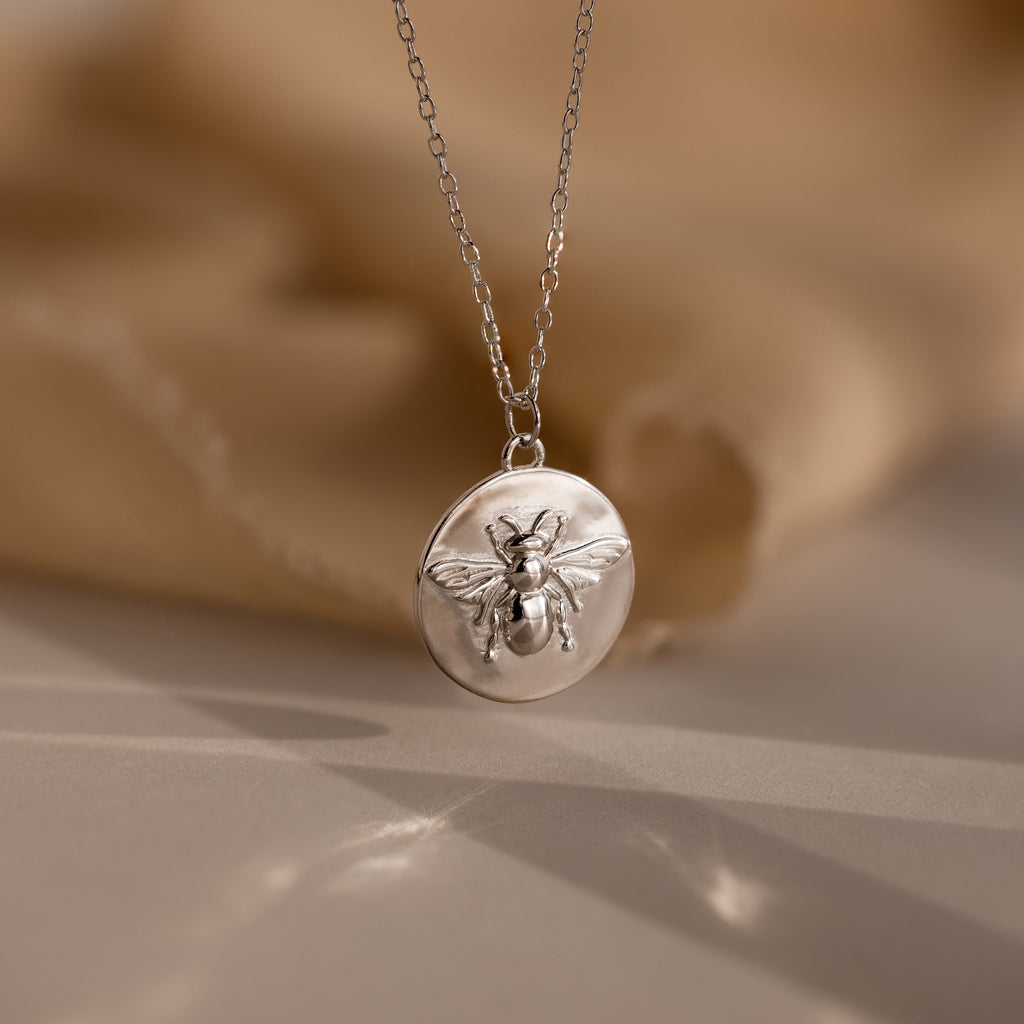 The Bee Coin Necklace features a silver coin pendant with a raised bee design, perfect for layering and shown on a soft beige background.