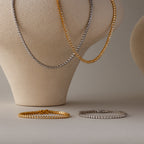 The Diamond Bezel Tennis Necklace & Bracelet Set, featuring gold and silver bracelet sets with bezel-set diamonds, is elegantly displayed on beige busts and a flat surface.