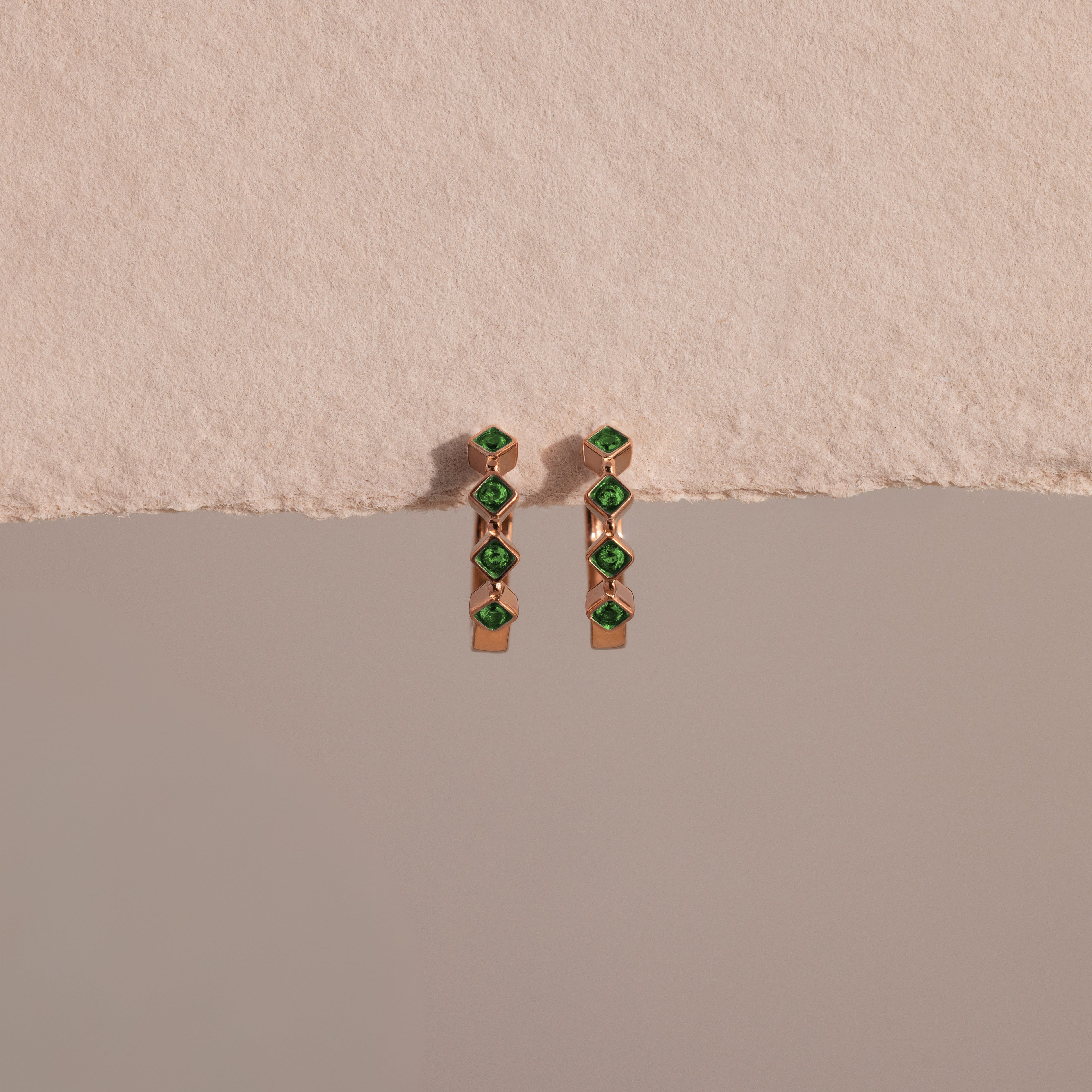 Geometric Emerald Huggies, gold earrings with five green gemstones each, are shown against a textured beige and smooth gray background.