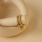 The Pave Triple Spike Huggies feature small clear stones in a bold, minimalist design. These striking hoop earrings are displayed on a curved beige surface.