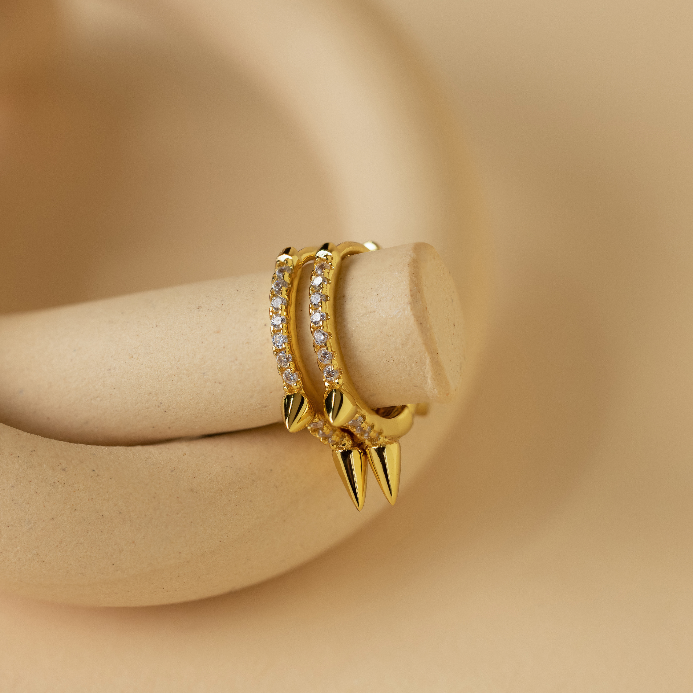 The Pave Triple Spike Huggies feature small clear stones in a bold, minimalist design. These striking hoop earrings are displayed on a curved beige surface.
