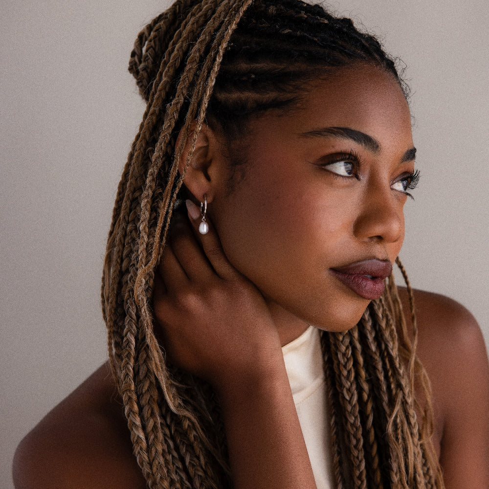 Woman with braided hair touching her face with silver pearl hoop earrings against a neutral background