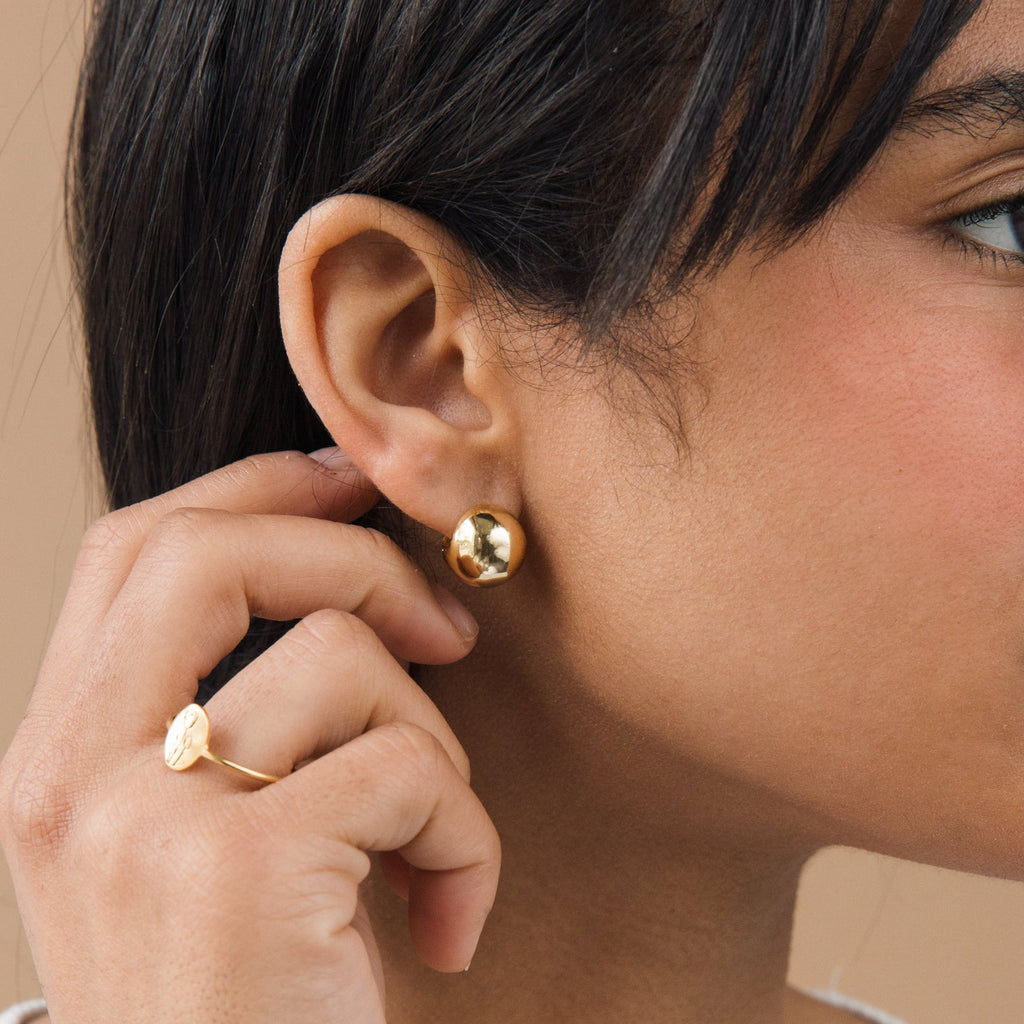 Close-up of a person wearing gold dome hoop earrings and a ring, with a neutral background.