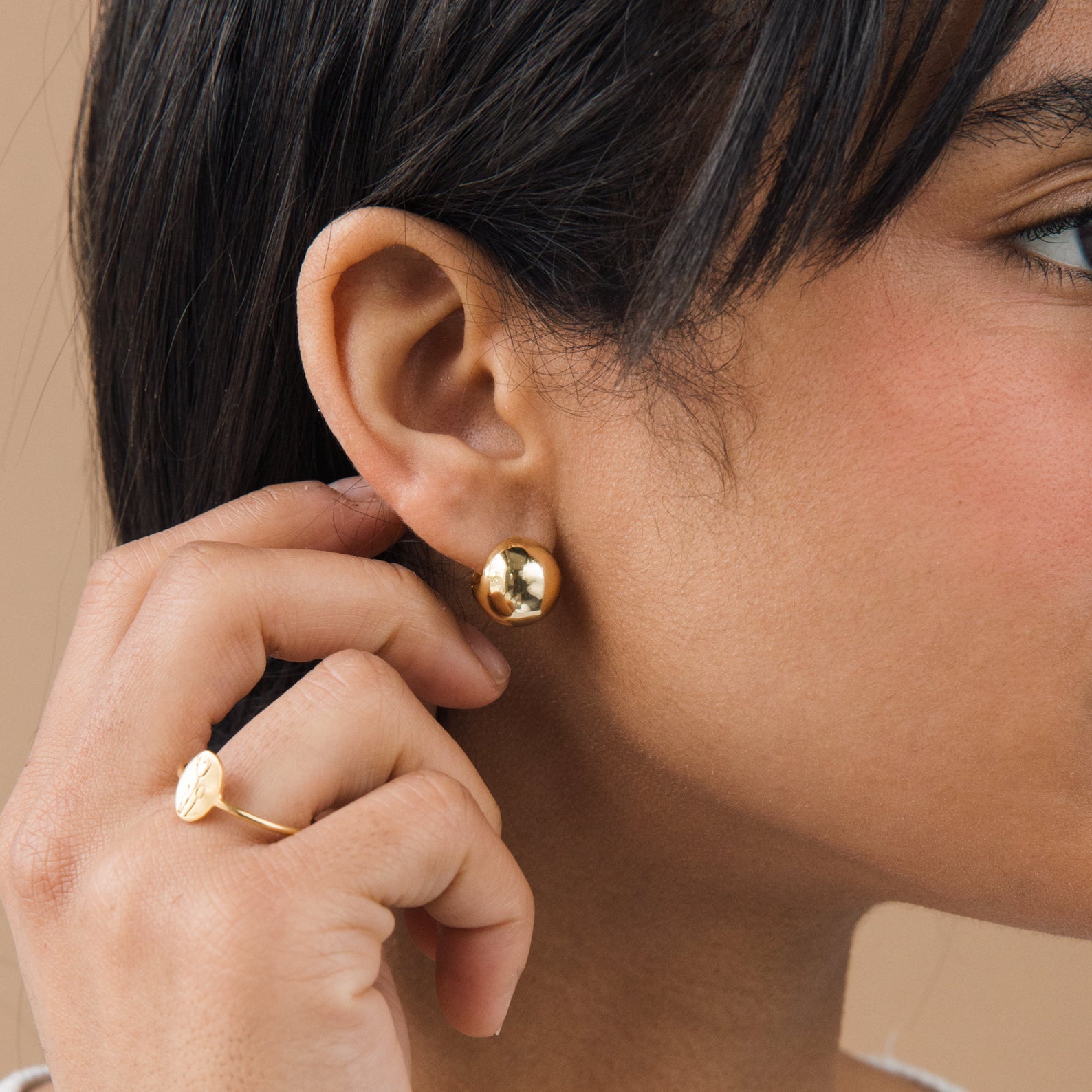 Close-up of a person wearing gold dome hoop earrings and a ring, with a neutral background.