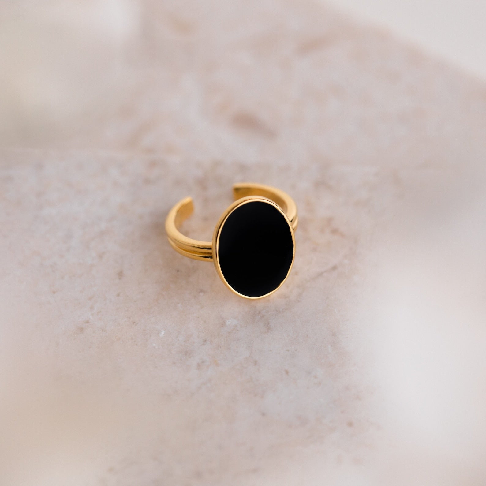 The Midnight Hour Onyx Ring features a large oval black stone and an adjustable band, elegantly displayed on a light beige marble surface.