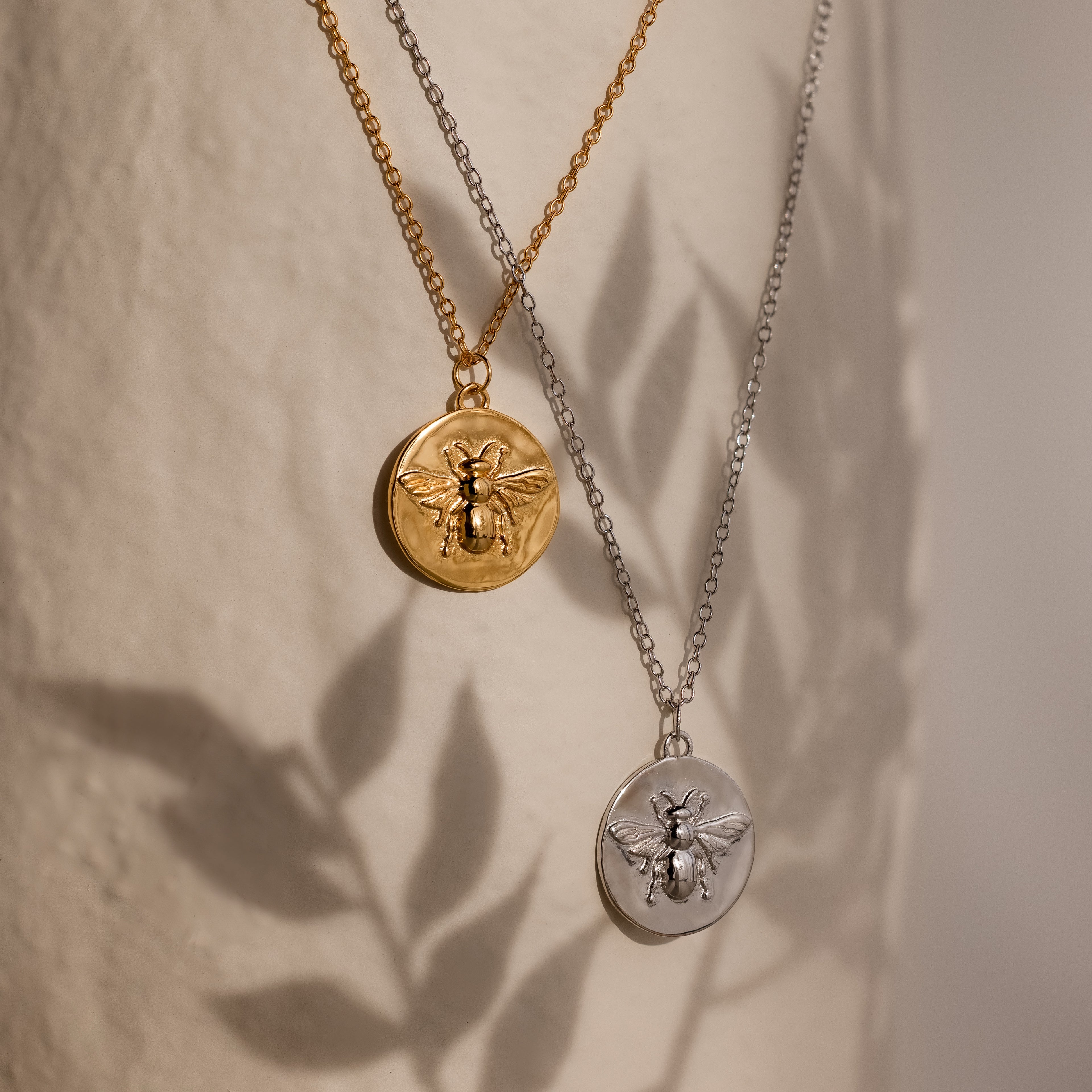 Two Bee Coin Necklaces, one gold and one silver, hang against a textured surface—perfect for layering and adding a chic honeybee accent to your style.