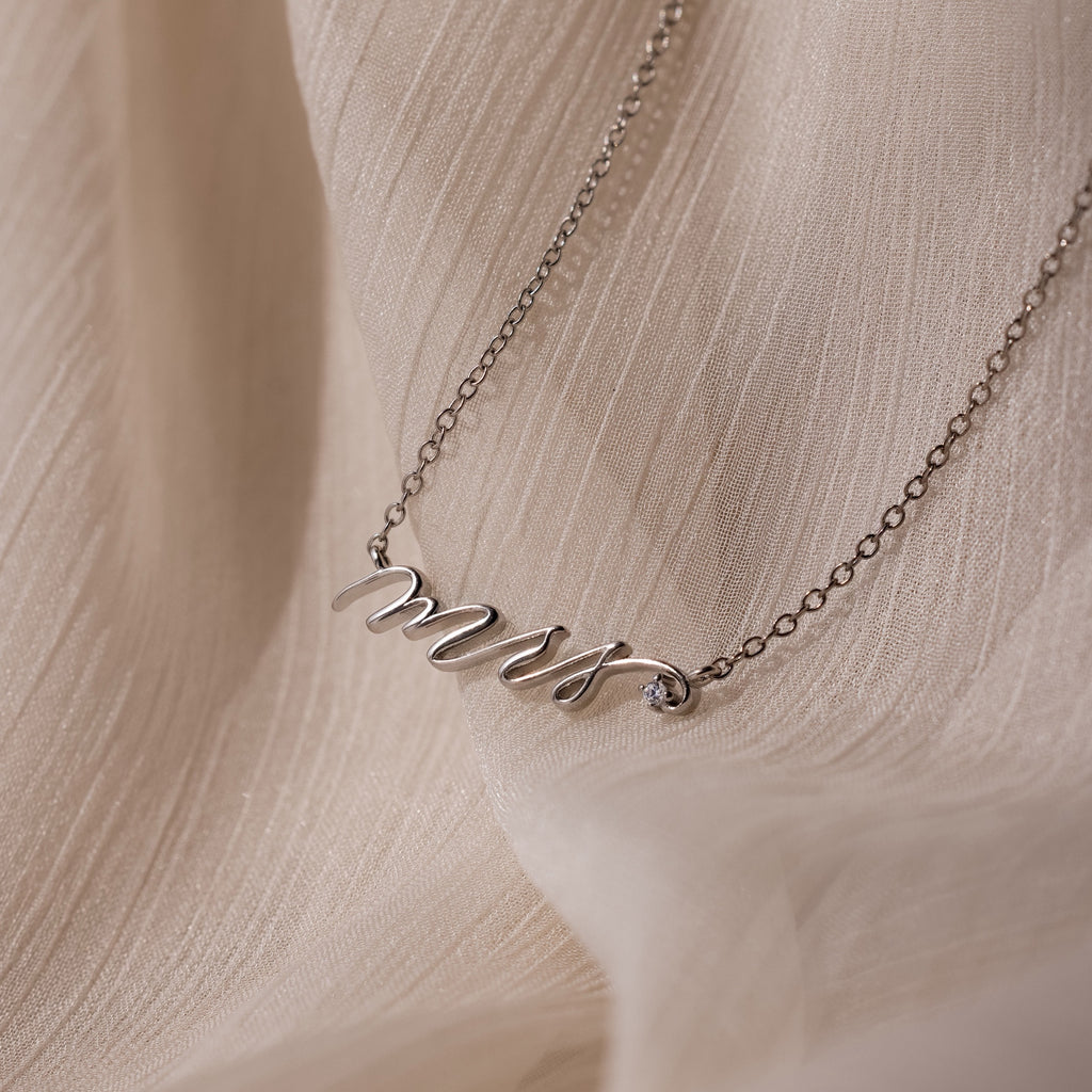 The Mrs. Necklace features a delicate script pendant, ideal as a wedding gift for any bride-to-be, showcased on a soft beige textured fabric background.