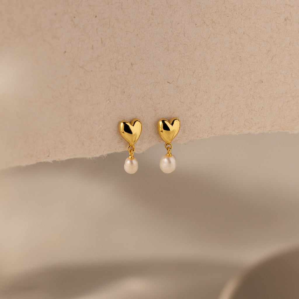 The Heart Pearl Drop Earrings feature gold heart-shaped studs with elegant pearl drops, shown on a neutral textured background.