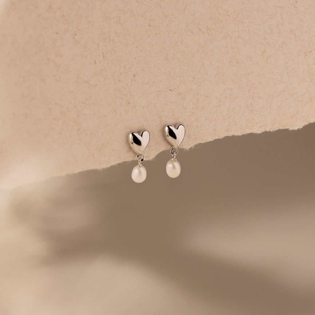 Heart Pearl Drop Earrings in Silver with heart-shaped studs and dangling pearls, elegantly displayed on a textured beige background.