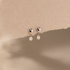 Heart Pearl Drop Earrings in Silver with heart-shaped studs and dangling pearls, elegantly displayed on a textured beige background.