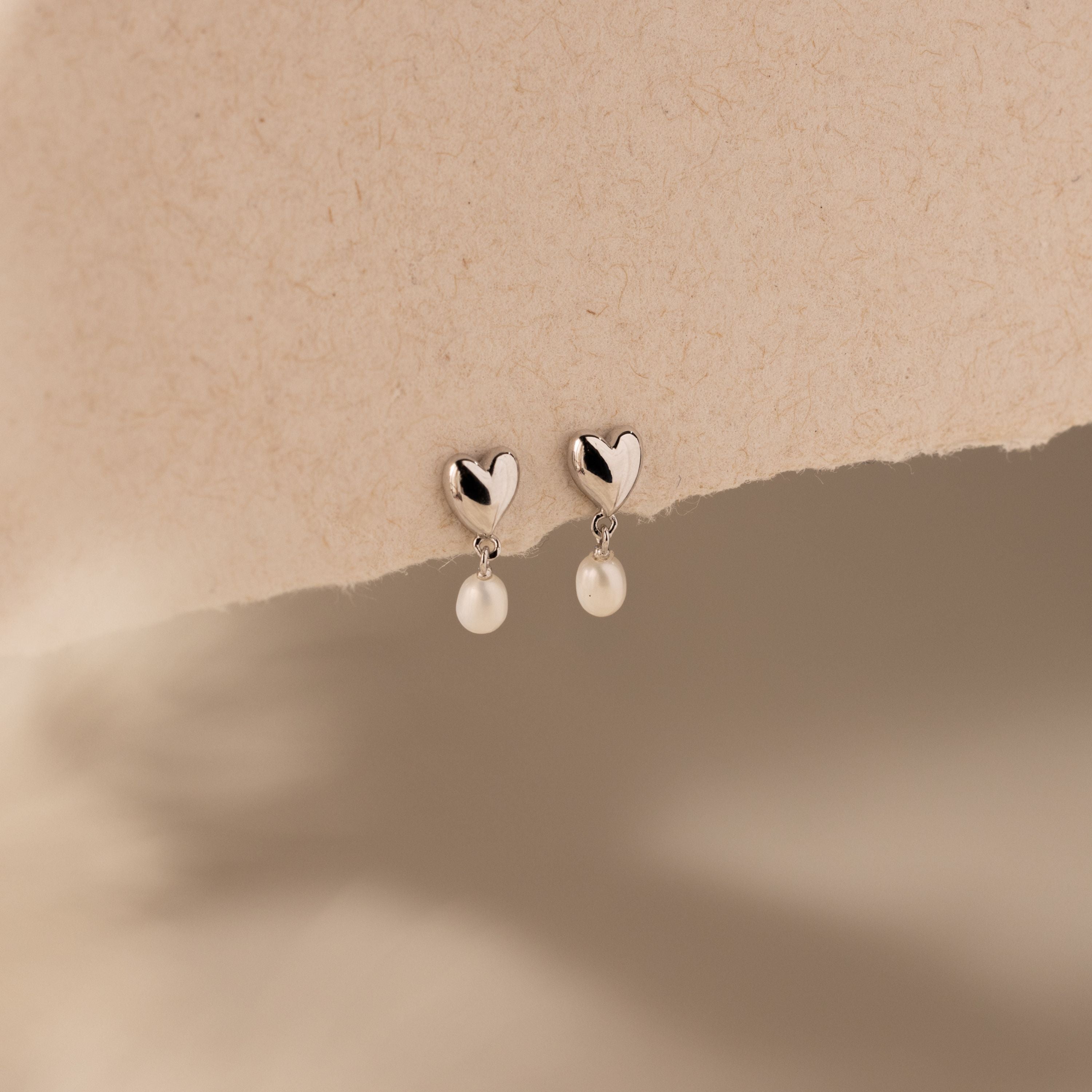 Heart Pearl Drop Earrings in Silver with heart-shaped studs and dangling pearls, elegantly displayed on a textured beige background.