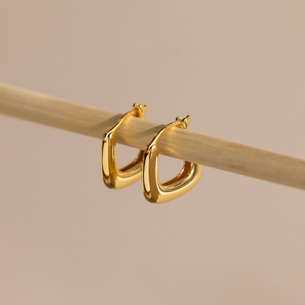 Close-up of gold geometric square hoop earrings hanging on a bamboo rod.