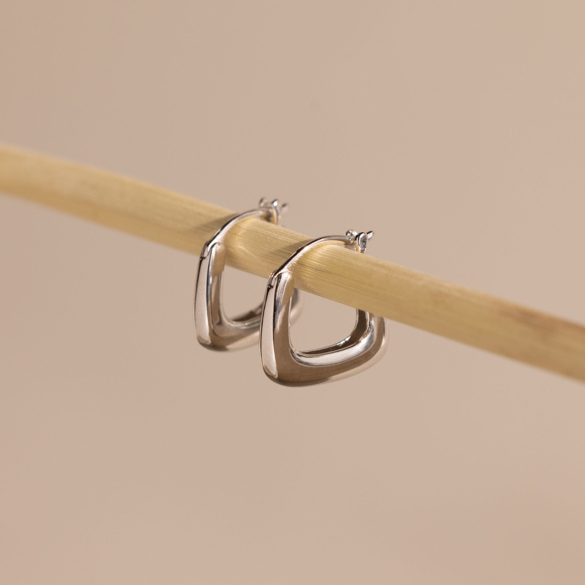 Pair of silver geometric square hoop earrings hanging on a wooden rod against a neutral background.