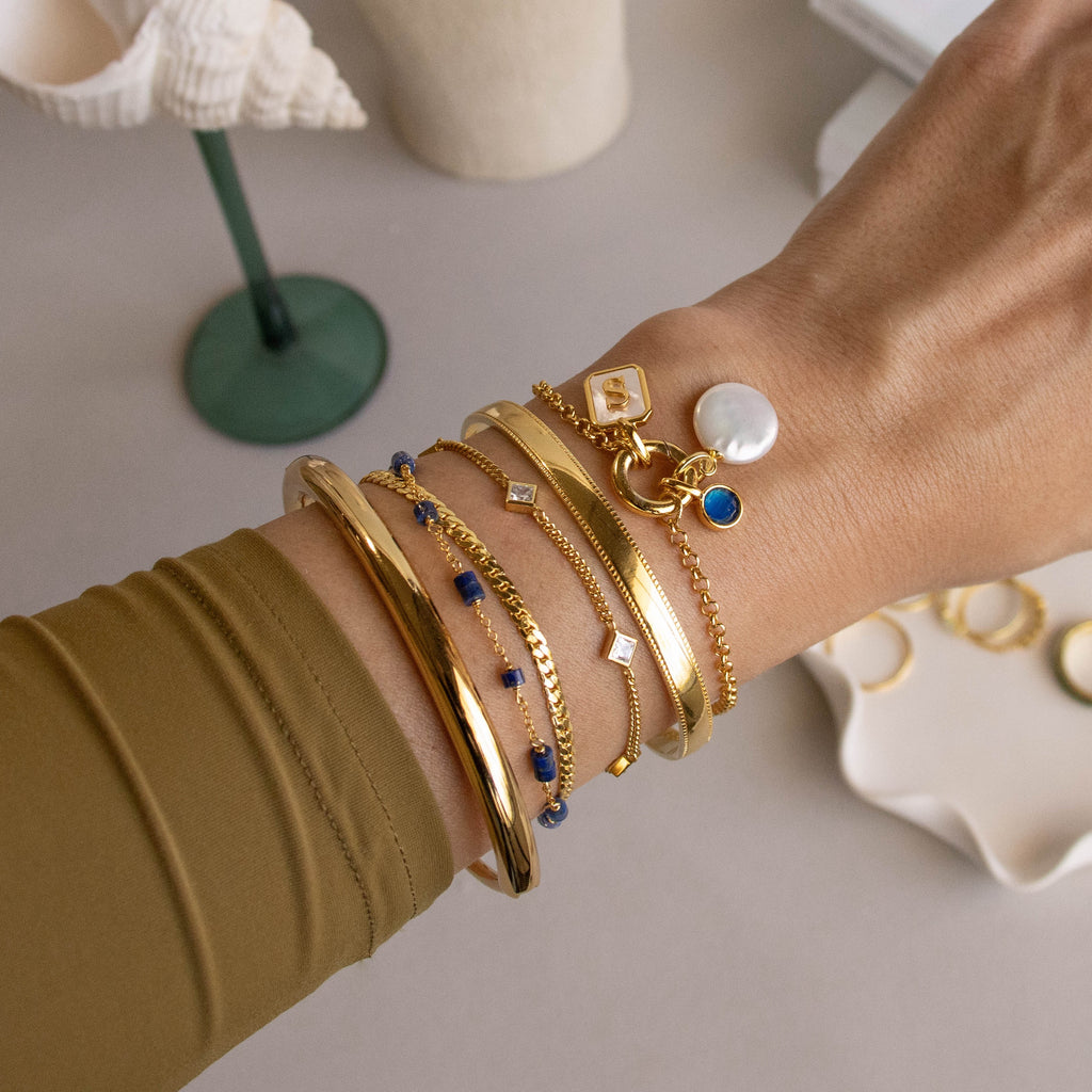 A hand showcases gold bangles, chain bracelets, and charms—like pearls and blue stones—styled with the Rolo Circle Charm Bracelet featuring a detachable hinged circle for adding charms.