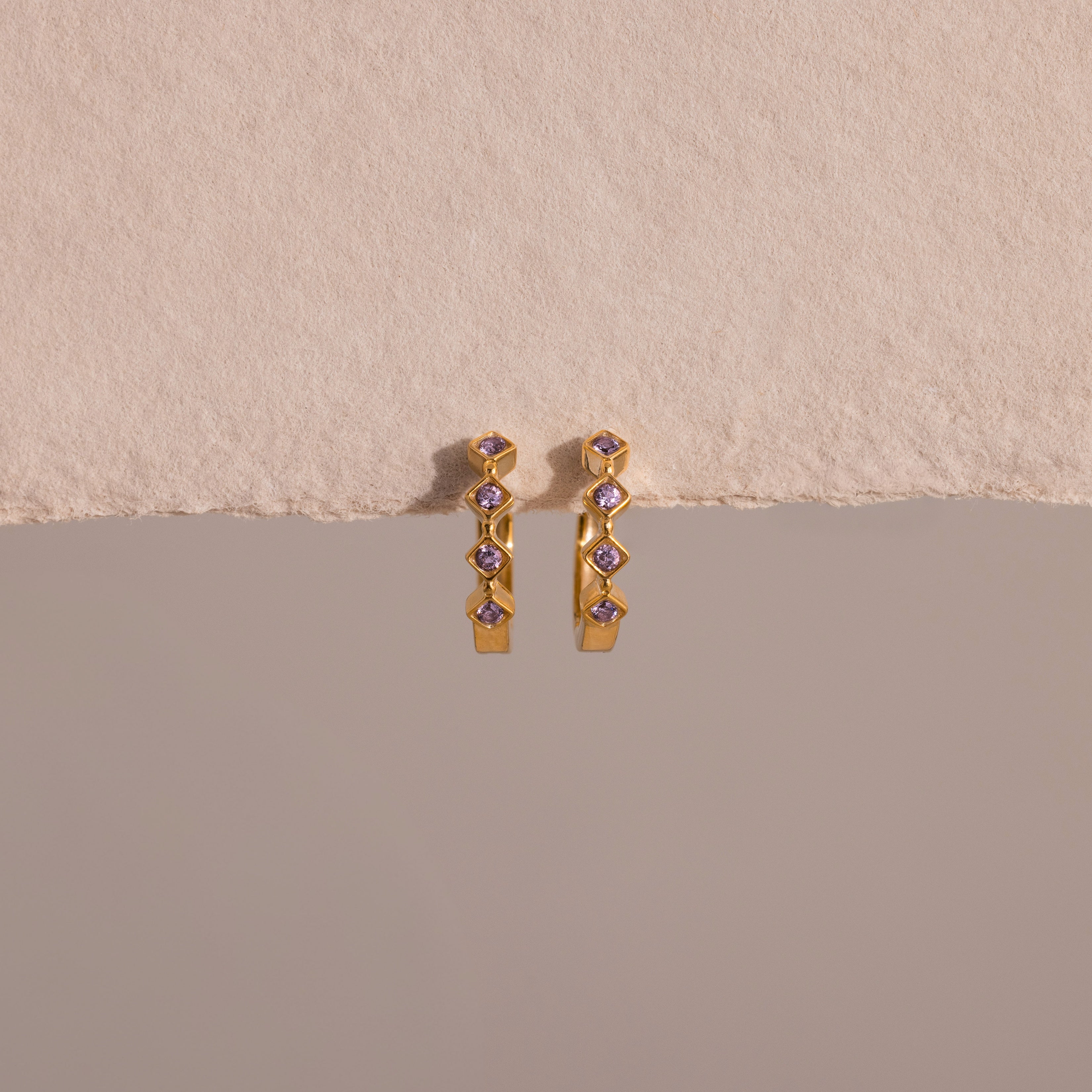 Geometric Alexandrite Huggies, gold earrings with geometric shapes and diamond accents, displayed on a textured beige surface.