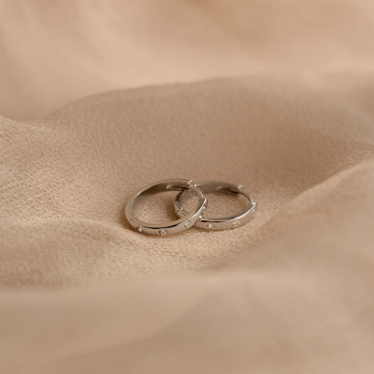 Two slim silver Classic Diamond Sprinkle Huggies with small clear stones set along the front, resting on draped beige fabric.