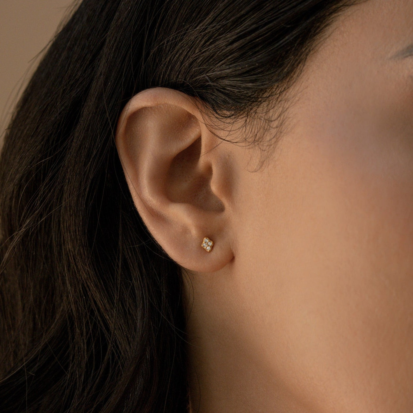 Close-up of a model wearing a gold diamond stud earring with dark hair and a beige background