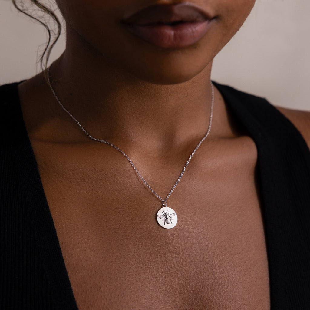A woman wearing a Bee Coin Necklace—a silver necklace featuring an engraved bee on a round charm, perfect for layering.