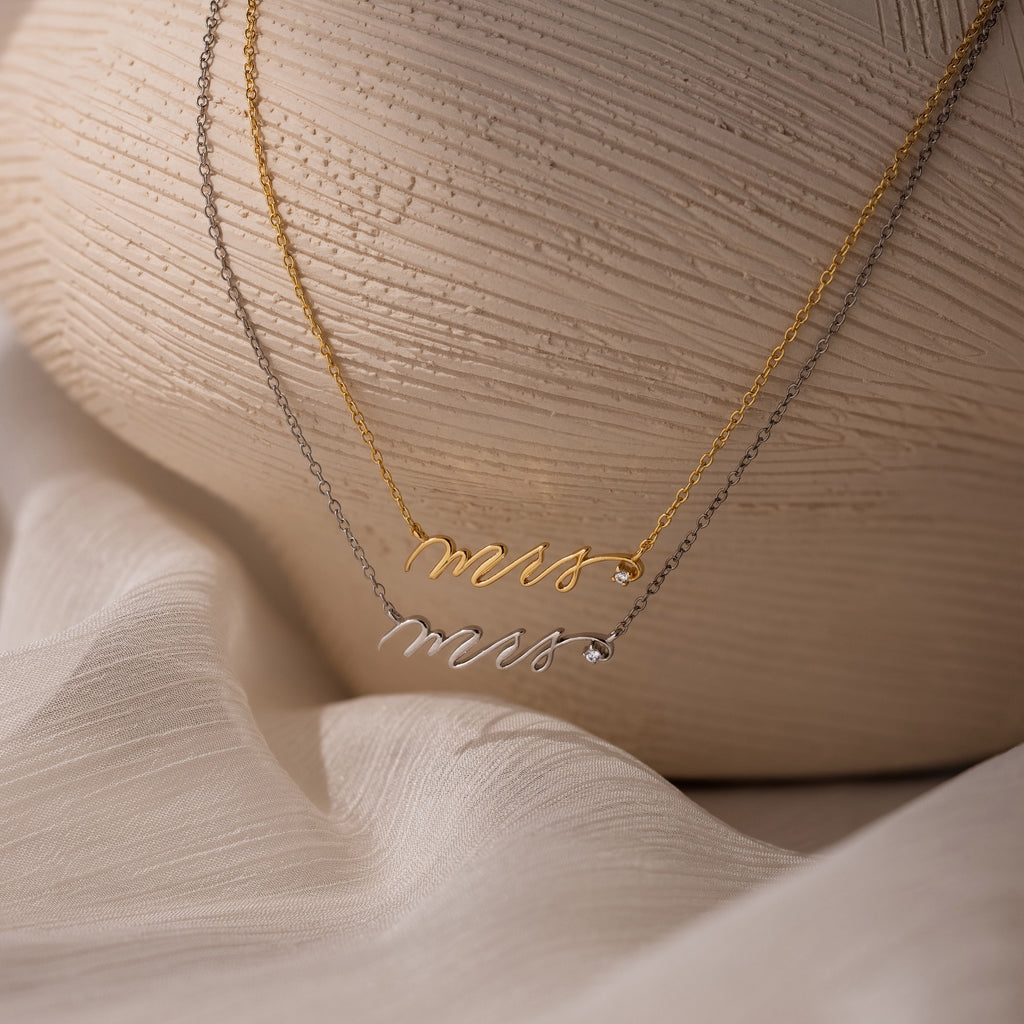 A gold and a silver script Mrs. necklace rest on a white textured surface—an ideal gift for the bride-to-be before she officially becomes a Mrs.