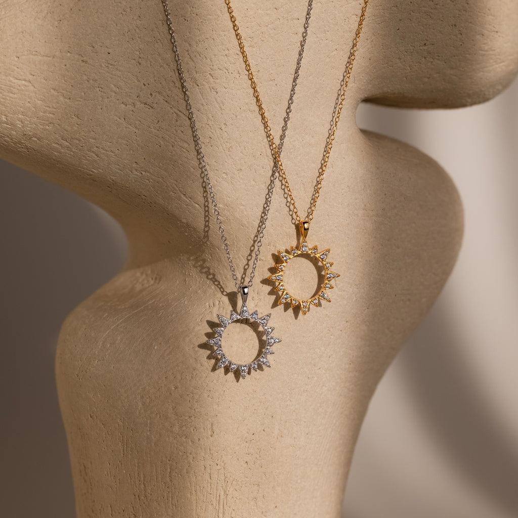 The Pave Sun Necklace—one silver with pave accents, one gold—shines on a textured beige mannequin bust, perfect for showcasing the collection.