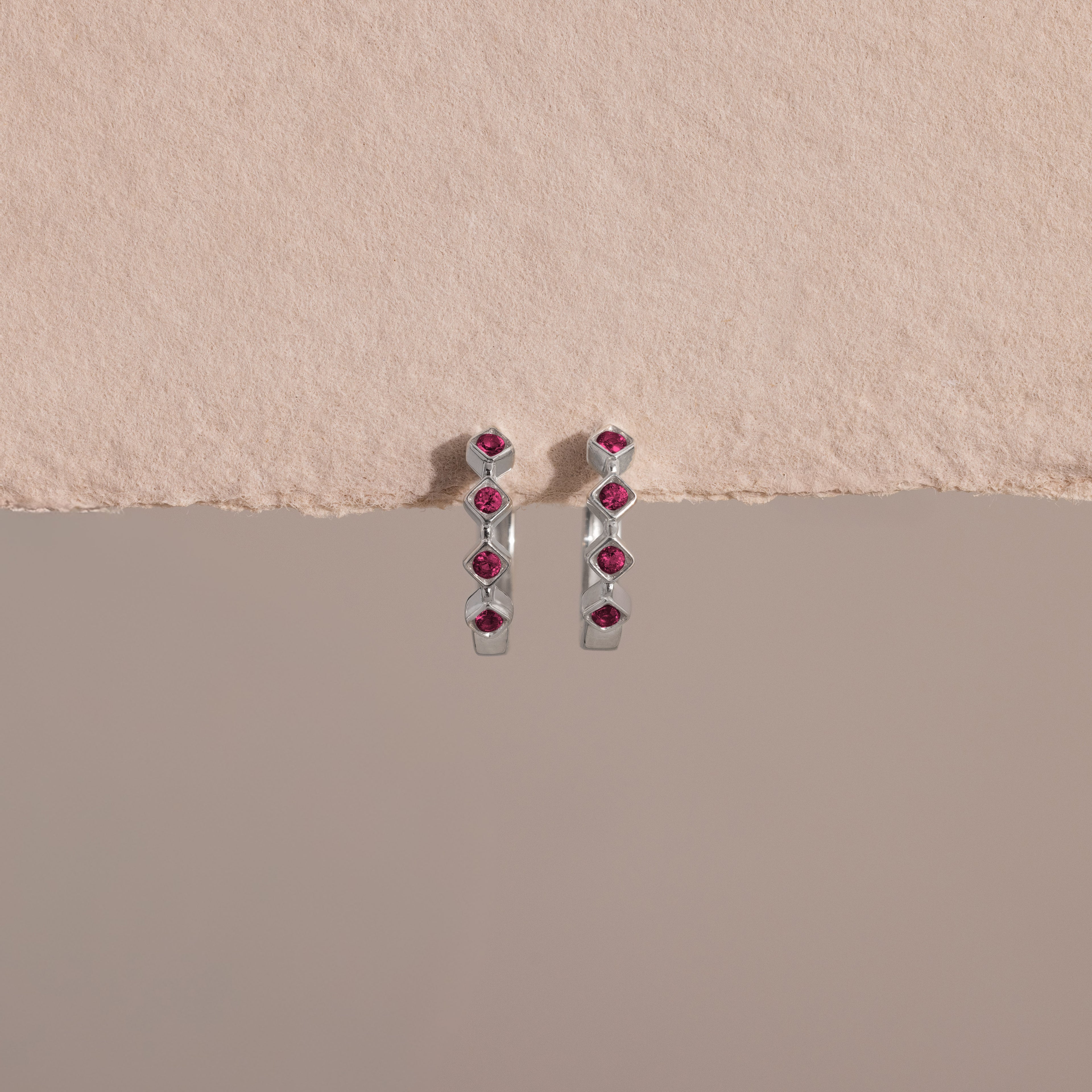 Geometric Ruby Huggies: silver earrings each set with six small red gemstones, offering elegant birthstone jewelry, displayed on a textured beige and gray background.