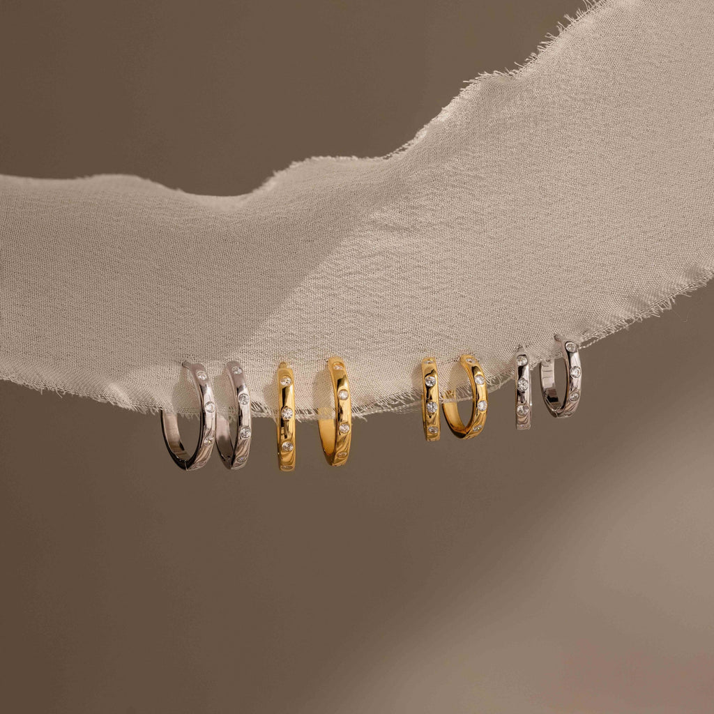 Six small hoop earrings in silver and gold tones displayed on a white textured band against a warm taupe background.
