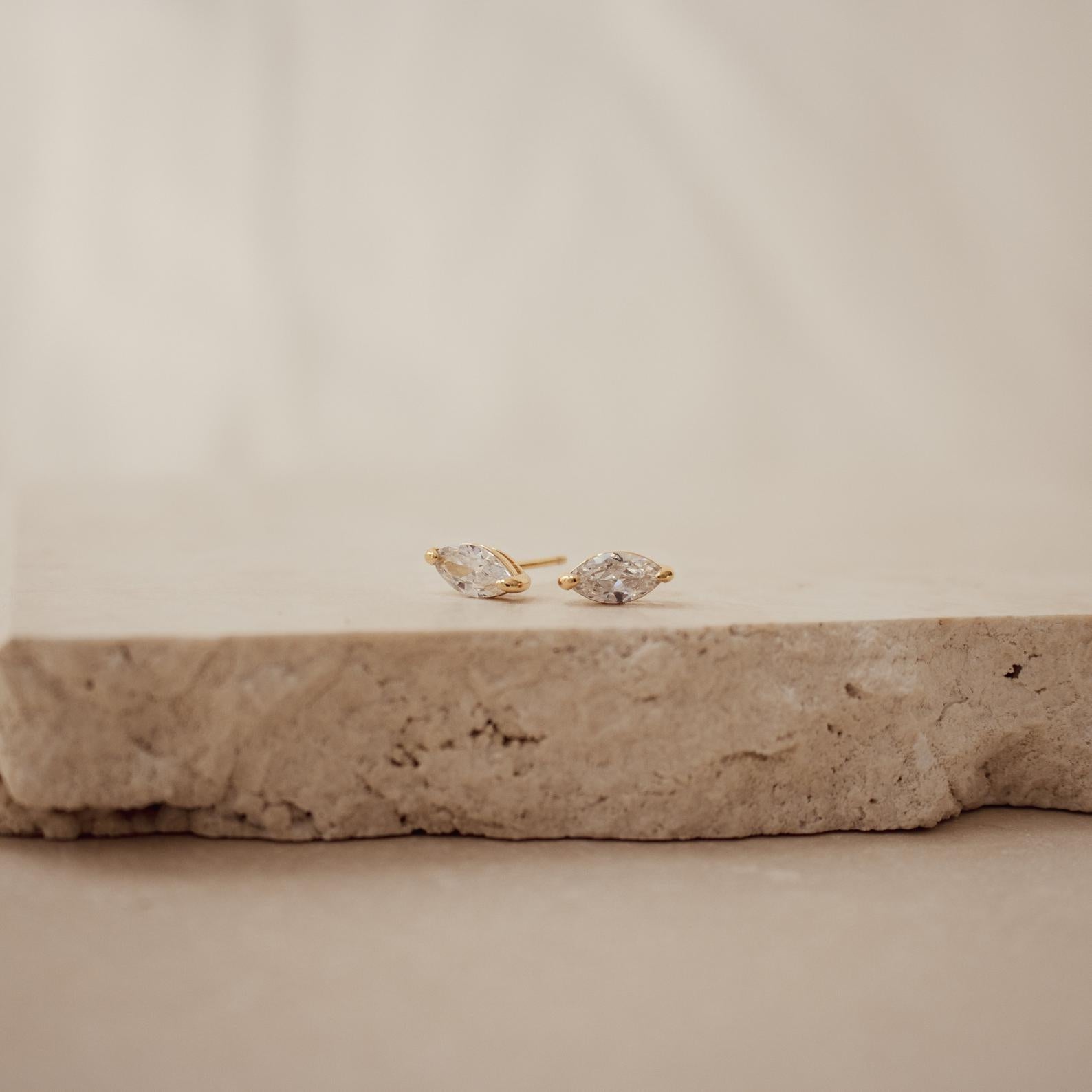 Pair of gold earrings with clear marquise shaped stones on a beige stone surface