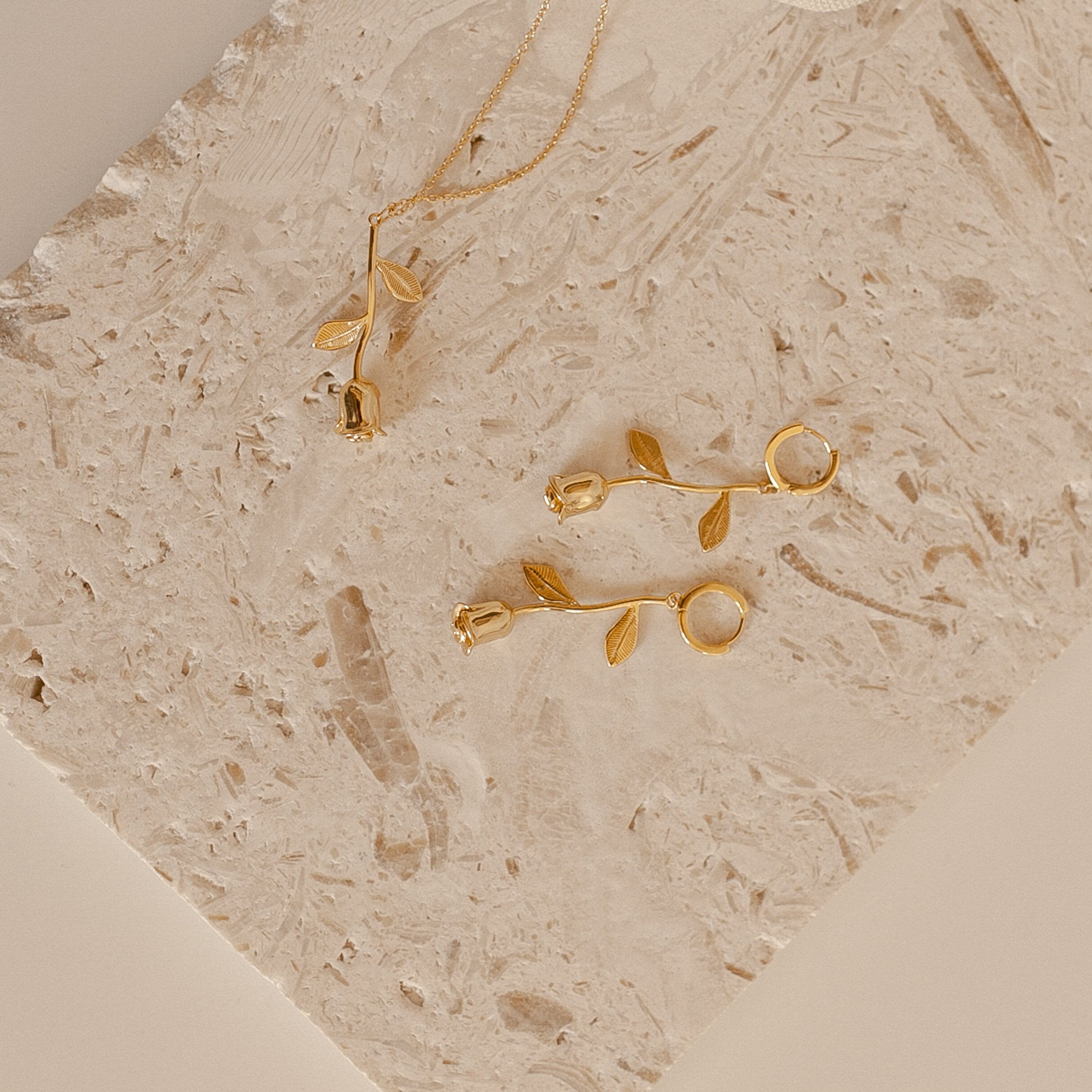 Gold rose necklace and rose earrings on a textured beige surface