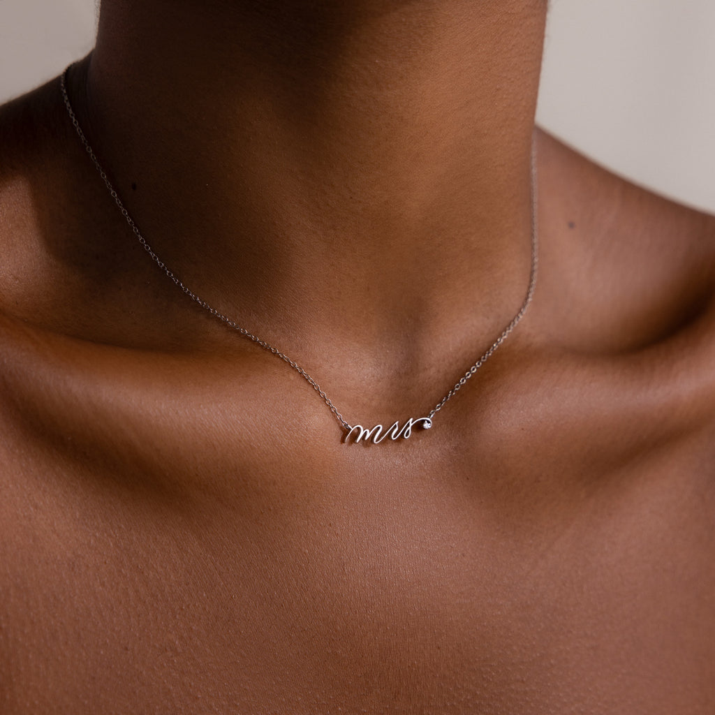 A Mrs. Necklace rests on a womans collarbone against brown skin, making it a perfect wedding gift for a bride-to-be.