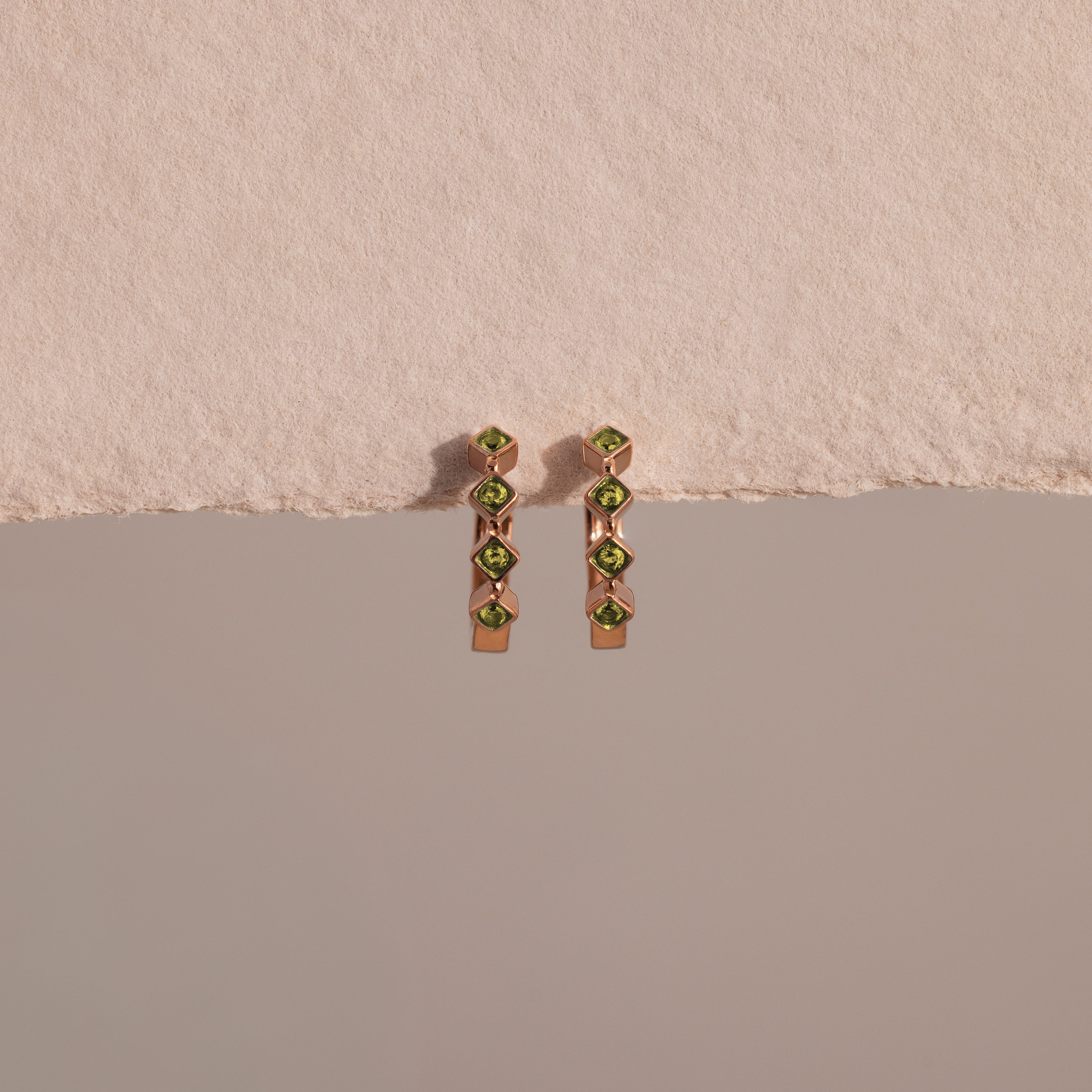 The Geometric Peridot Huggies, featuring green gemstones and diamond accents, are displayed against a soft pink textured background.