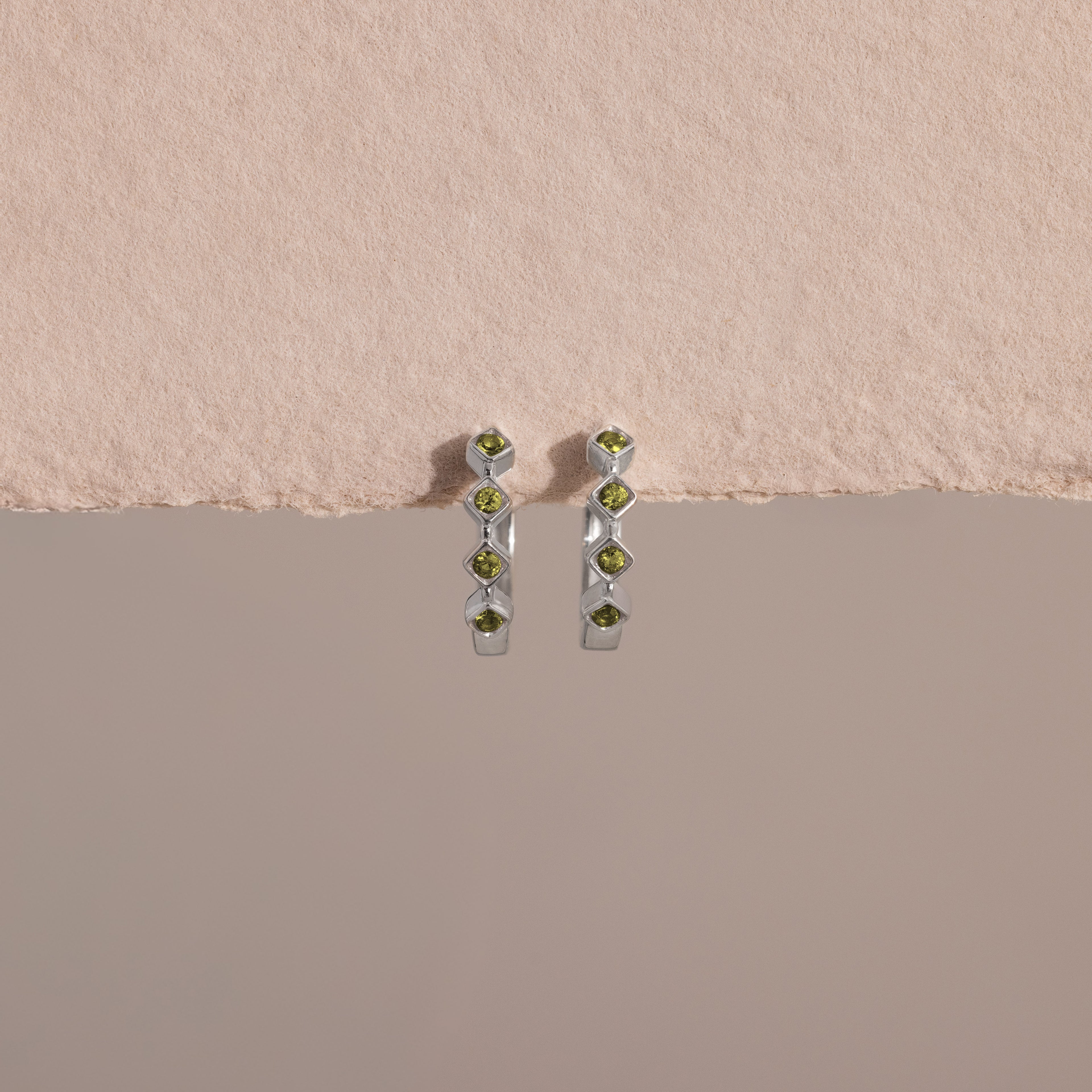 Geometric Peridot Huggies, each adorned with five green gemstones and diamond accents, are shown against a textured beige and smooth gray background.
