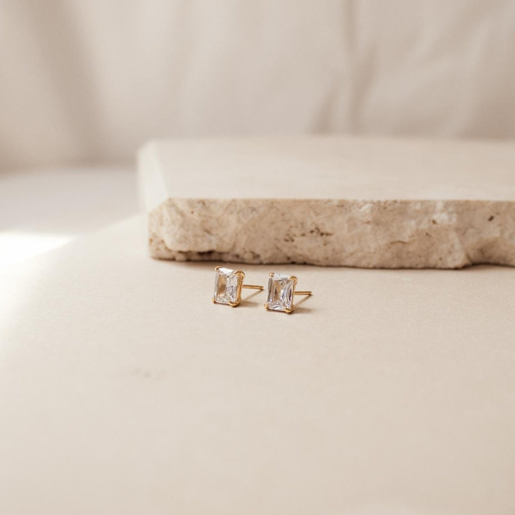 Gold stud earrings with emerald cut diamonds on a beige surface with a neutral background